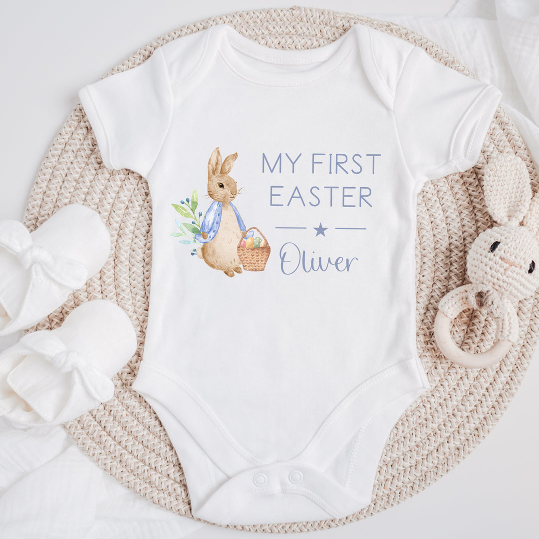 Personalised white Easter baby vest that says 'My First Easter Oliver'. This design features a rabbit wearing a blue jacket holding an Easter basket full of eggs