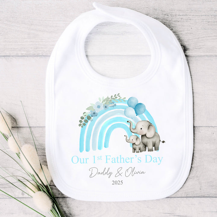 Personalised white Father's Day baby bib that says 'Our 1st Father's Day Daddy & Olivia 2025'. This design features a blue rainbow with 2 grey elephants holding 3 balloons