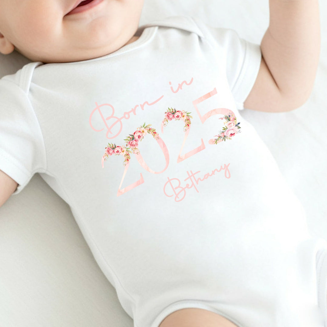 a personalised white baby vest that says 'Born in 2025 Bethany'. This design features a pink floral elements around the 2025