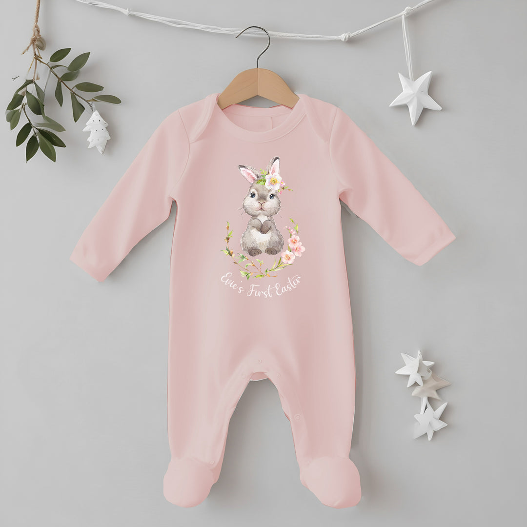 Personalised pink Easter baby grow/sleepsuit that says 'Evie's First Easter'. This design features a bunny in a pink floral wreath