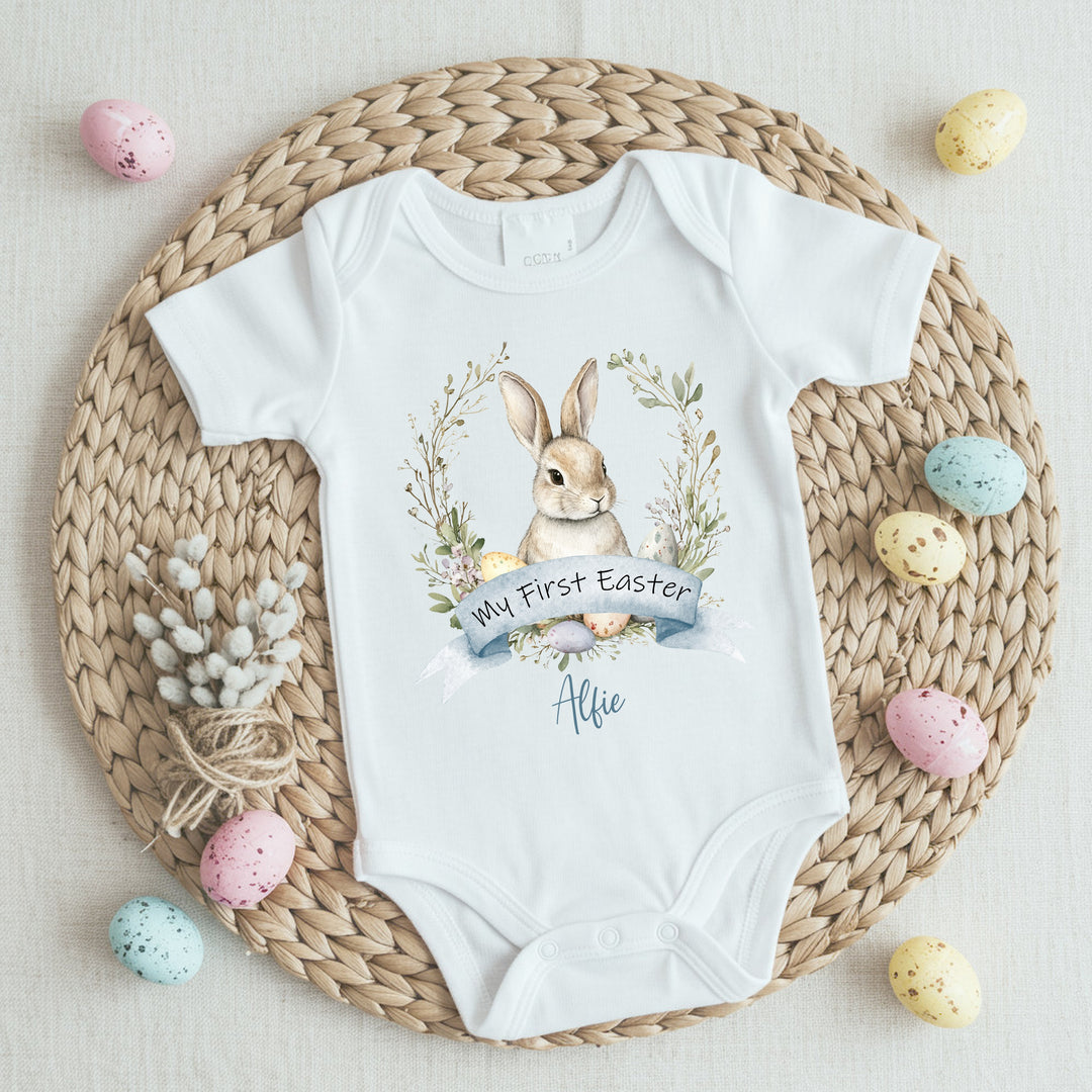 Personalised white Easter baby vest that says 'My First Easter Alfie'. This design features a brown bunny in a wreath with Easter eggs