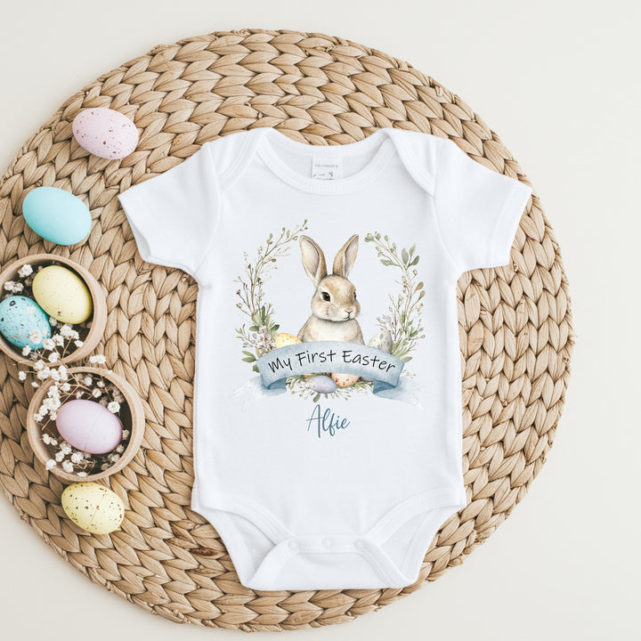 Personalised white Easter baby vest that says 'My First Easter Alfie'. This design features a brown bunny in a wreath with Easter eggs