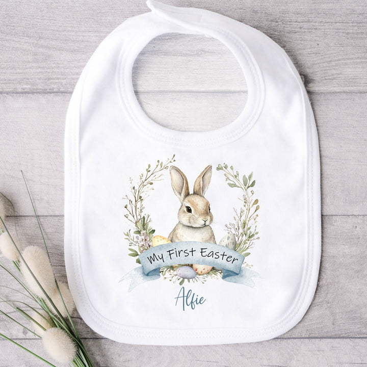 Personalised white Easter baby bib that says 'My First Easter Alfie'. This design features a brown bunny in a wreath with Easter eggs