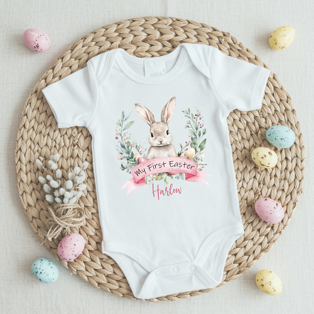 Personalised white Easter baby vest that says 'My First Easter Harlow'. This design features a white bunny in the middle of a wreath with Easter eggs at the bottom of the wreath