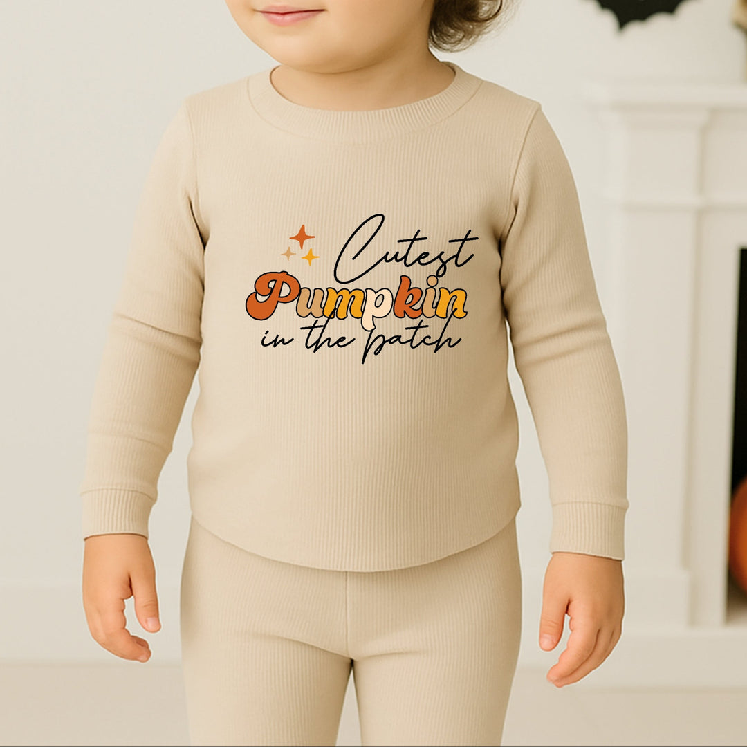 Child wearing a beige outfit with 'Cutest Pumpkin in the Patch' text, standing in front of Halloween decorations.