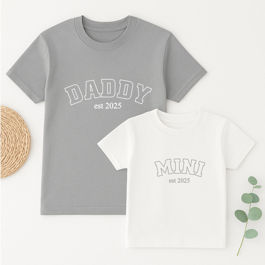 Gray t-shirt with 'DADDY est 2025' text and white t-shirt with 'MINI est 2025' text on a light background.