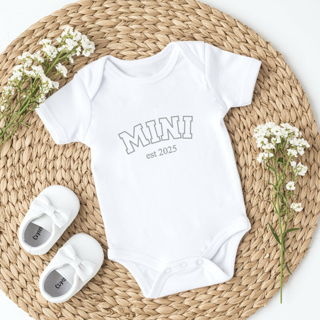 White baby onesie with 'MINI est 2025' text, white baby shoes, and baby's breath flowers on a woven mat.