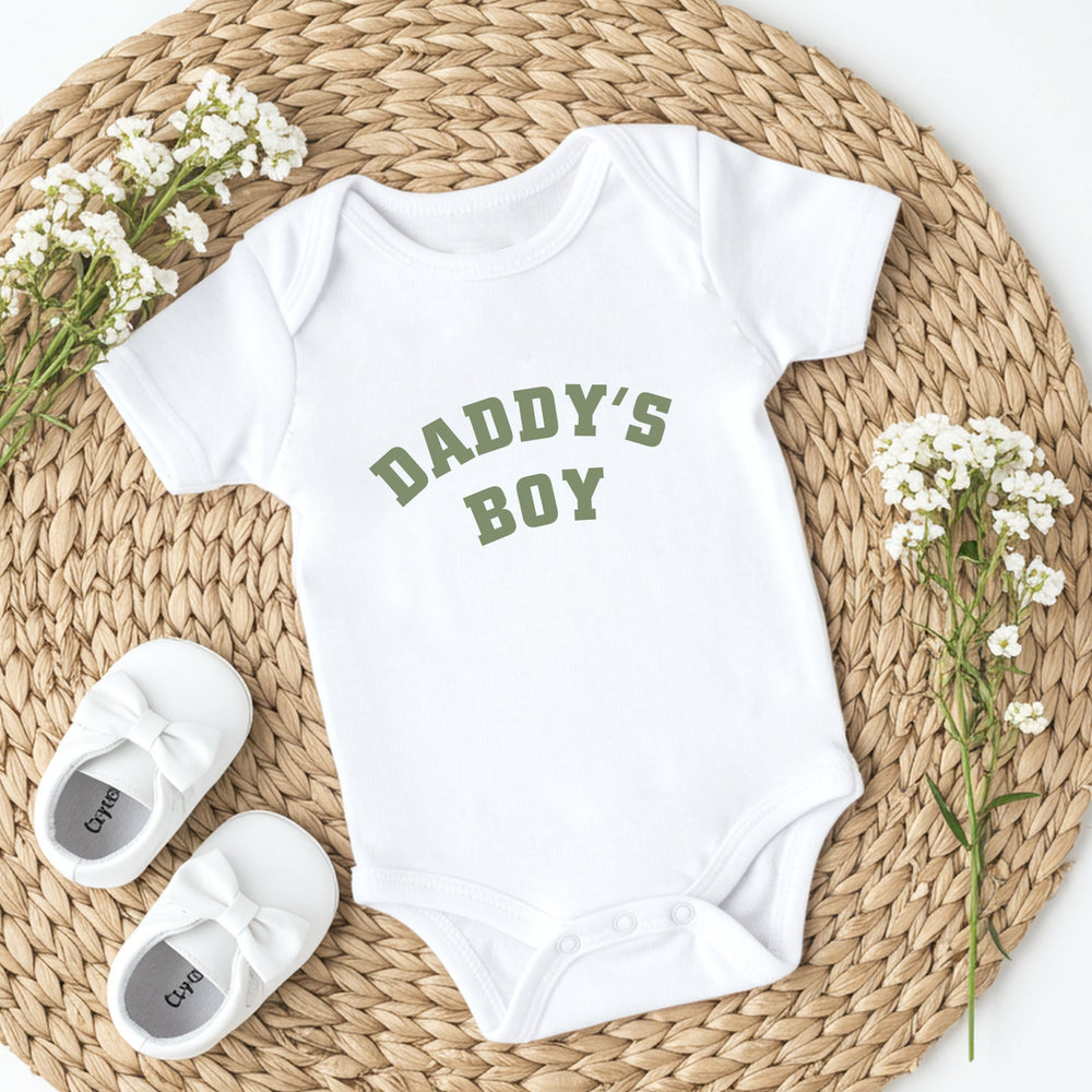 White baby onesie with 'Daddy's Boy' text on a woven mat with baby shoes and flowers.