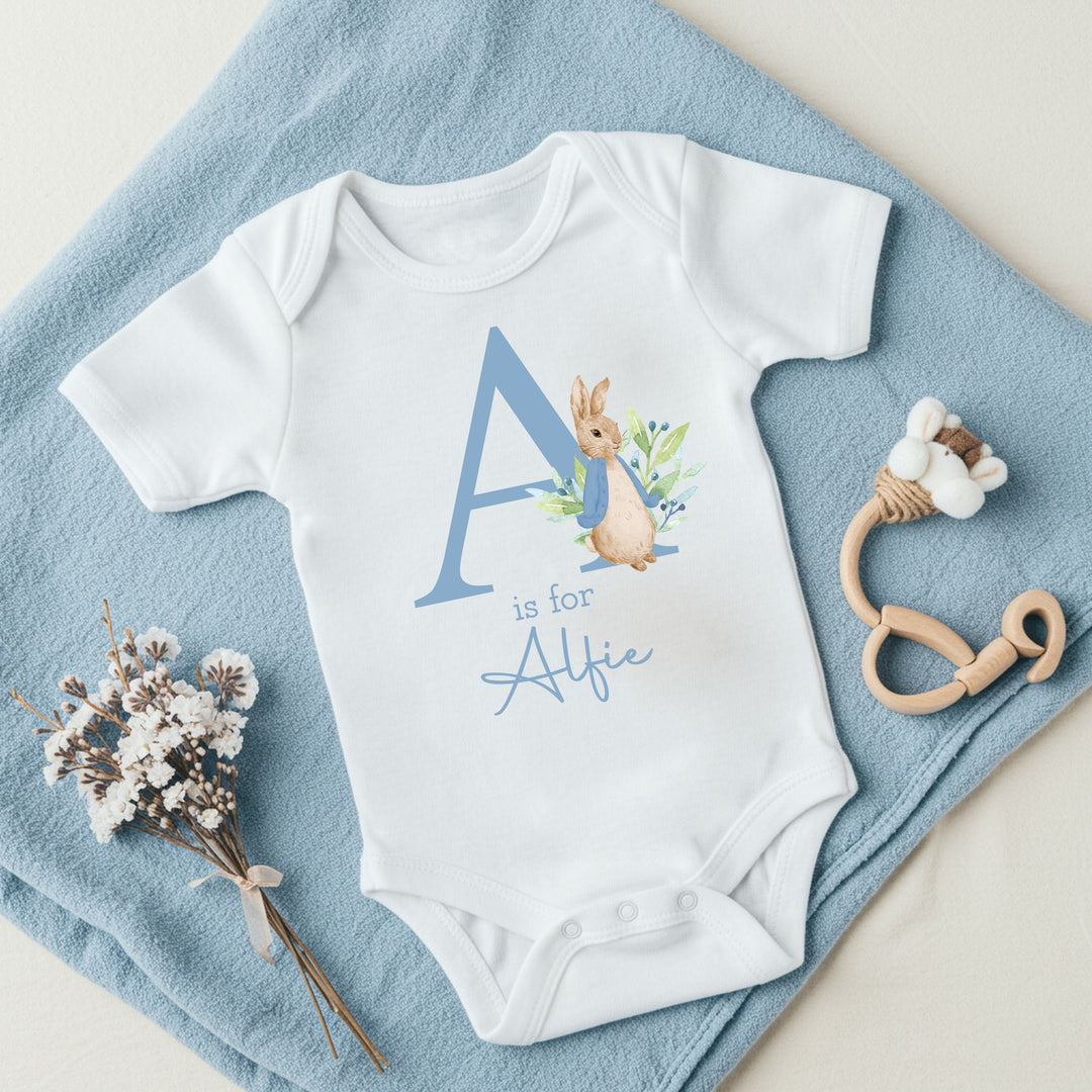 a personalised white baby vest that says 'A is for Alfie' in dusky blue text. This design features a rabbit wearing a dusky blue jacket