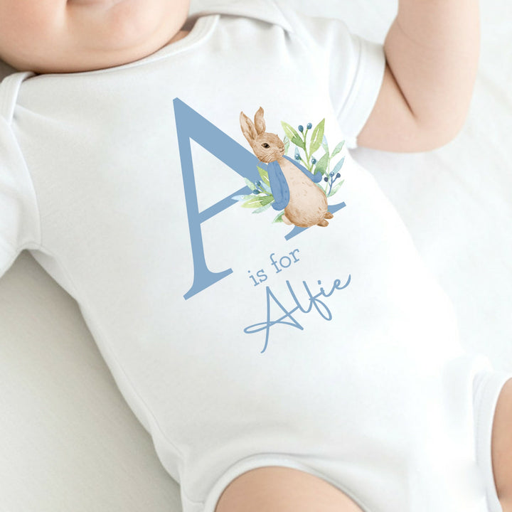 a personalised white baby vest that says 'A is for Alfie' in dusky blue text. This design features a rabbit wearing a dusky blue jacket