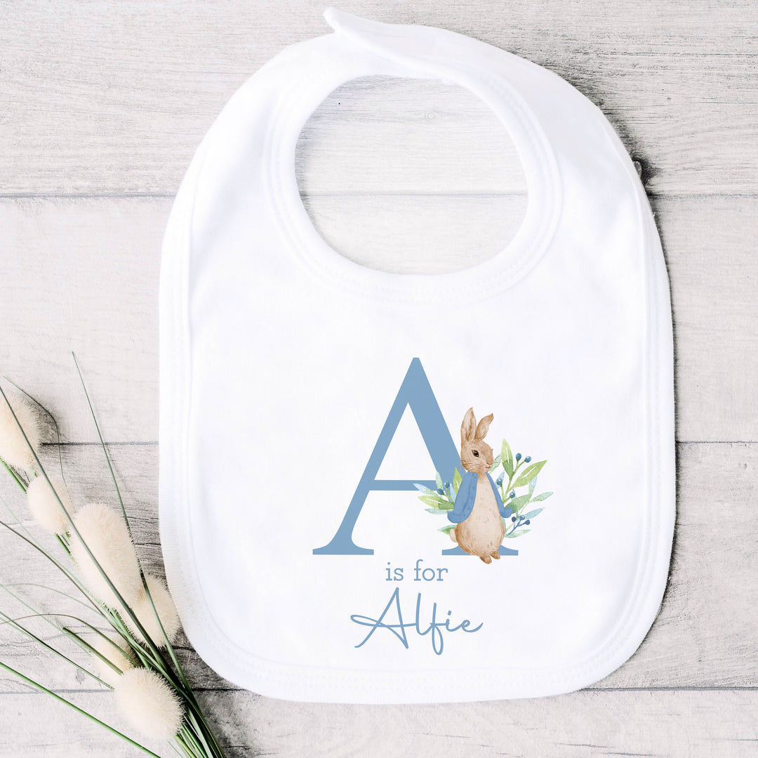 a personalised white baby bib that says 'A is for Alfie' in dusky blue text. This design features a rabbit wearing a dusky blue jacket