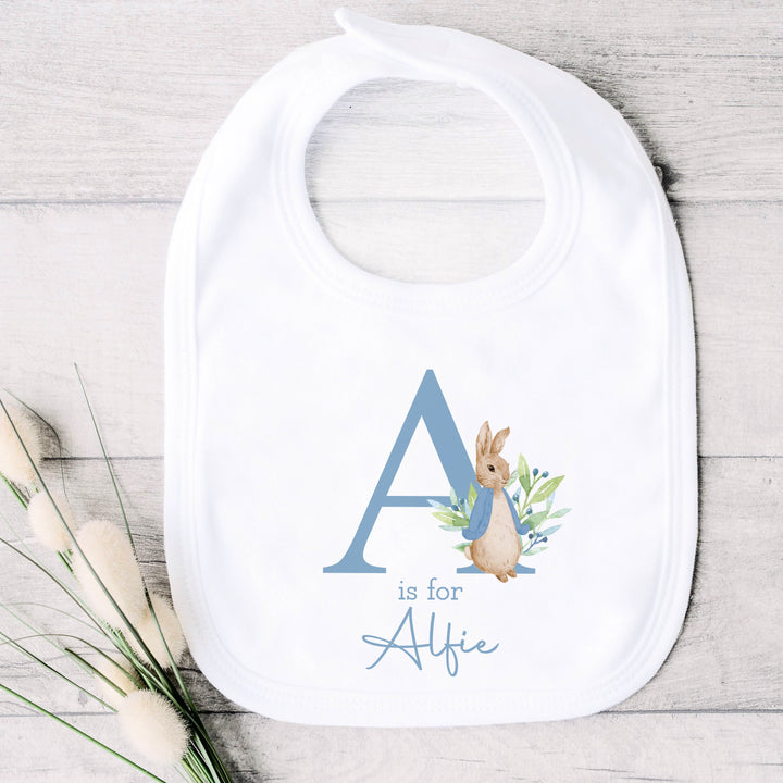 a personalised white baby bib that says 'A is for Alfie' in dusky blue text. This design features a rabbit wearing a dusky blue jacket