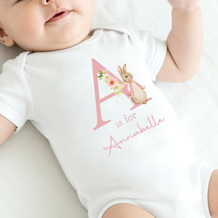 A personalised white baby vest that says 'A is for Annabelle' in pink. This design features a rabbit wearing a pink jacket