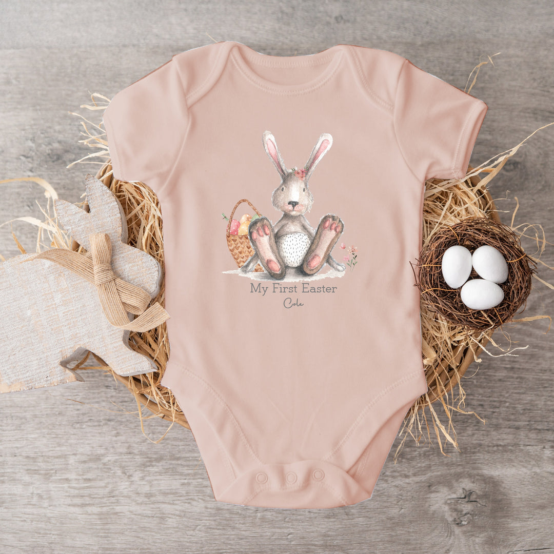 Personalised Pink Easter baby vest that says 'My First Easter Cole'. This design features a grey rabbit with pink details sitting next to an Easter basket with eggs in it 