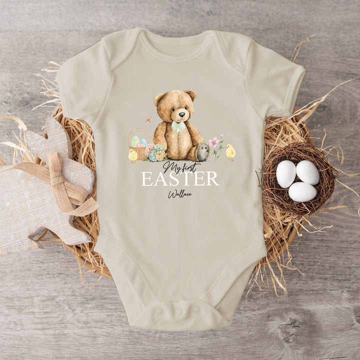 Peronalised beige Easter baby vest that says 'My First Easter Wallace'. This design features a teddy bear wearing a sage green bow tie sitting with an Easter basket and 2 chicks