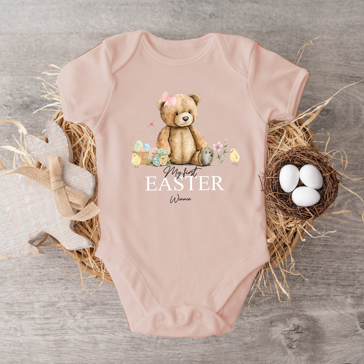 Personalised pink Easter baby vest that says 'My First Easter Winnie'. This design features a teddy bear wearing a bow sitting next to an Easter basket and 2 yellow chicks