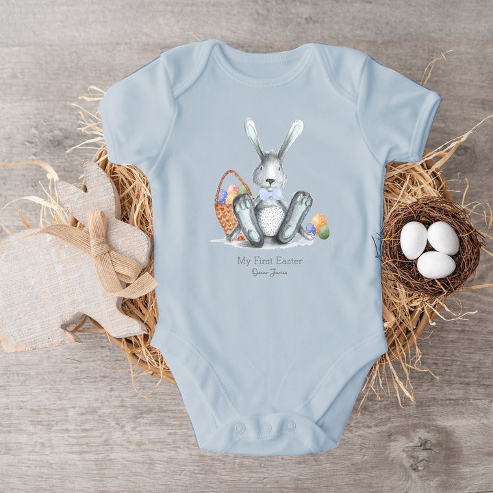 Personalised blue Easter baby vest that says 'My First Easter Oscar James'. This design features a grey rabbit with blue details sitting next to an Easter basket full of eggs