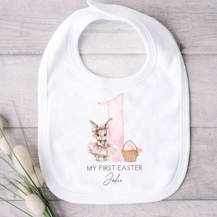 Personalised white Easter baby bib that says 'My First Easter Jodie'. This design features a big pink 1 with a ballerina bunny one side and an Easter basket with pink Easter eggs in it the other side