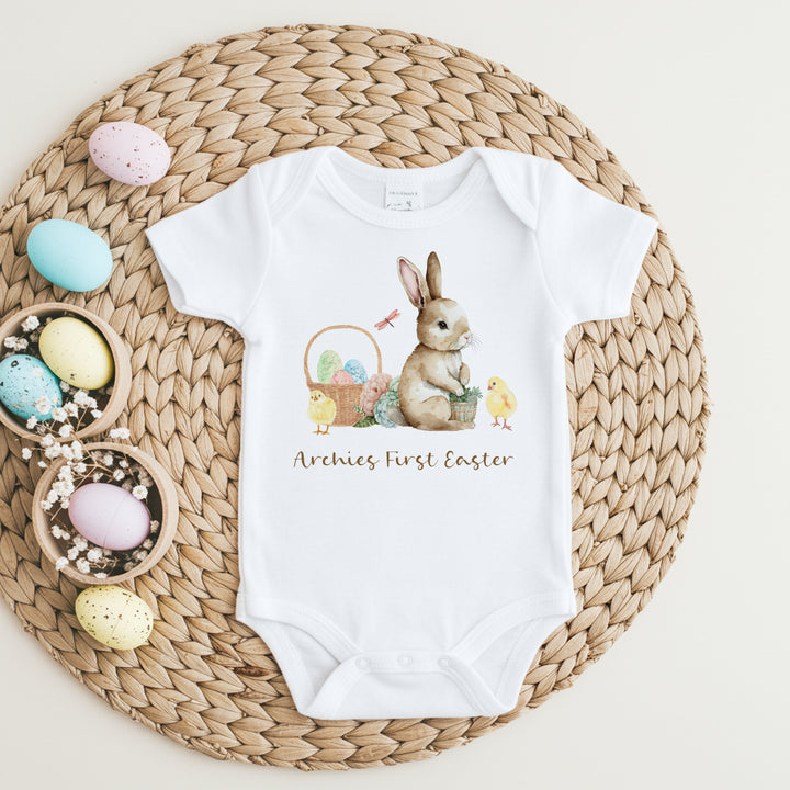 Personalised white Easter baby vest that says 'Archie's First Easter'. This design features a brown rabbit sitting next to an Easter basket and 2 yellow chicks