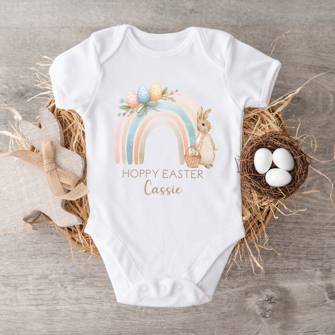 Personalised white Easter baby vest that says 'Hoppy Easter Cassie'. This design features a pastel rainbow with a rabbit wearing a beige jacket holding an Easter basket next to it