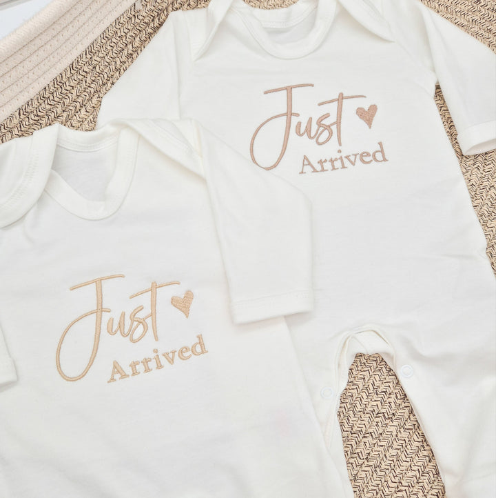 This photo shows 2 white babygrows that have 'Just Arrived' embroidered on them. This design features a small heart and shows 2 of the avaliable font colours (Sand and Fawn)