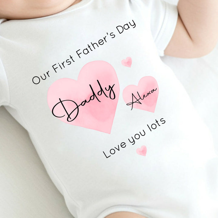 Personalised white Father's Day baby vest that says 'Our First Father's Day Daddy Alexa Love you lots'. This design features 2 big pink hearts