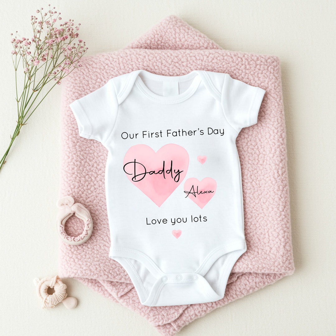 Personalised white Father's Day baby vest that says 'Our First Father's Day Daddy Alexa Love you lots'. This design features 2 big pink hearts