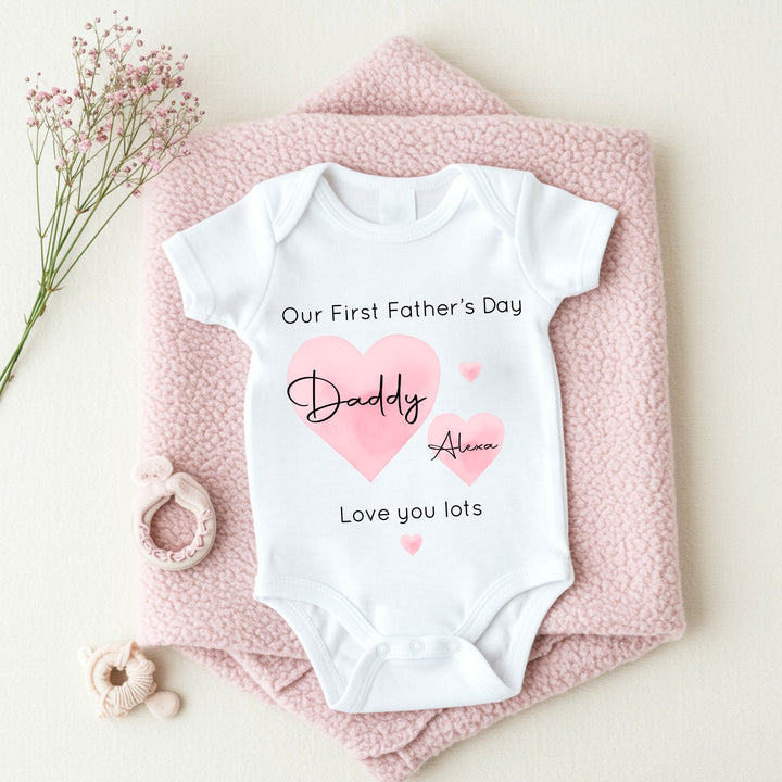 Personalised white Father's Day baby vest that says 'Our First Father's Day Daddy Alexa Love you lots'. This design features 2 big pink hearts