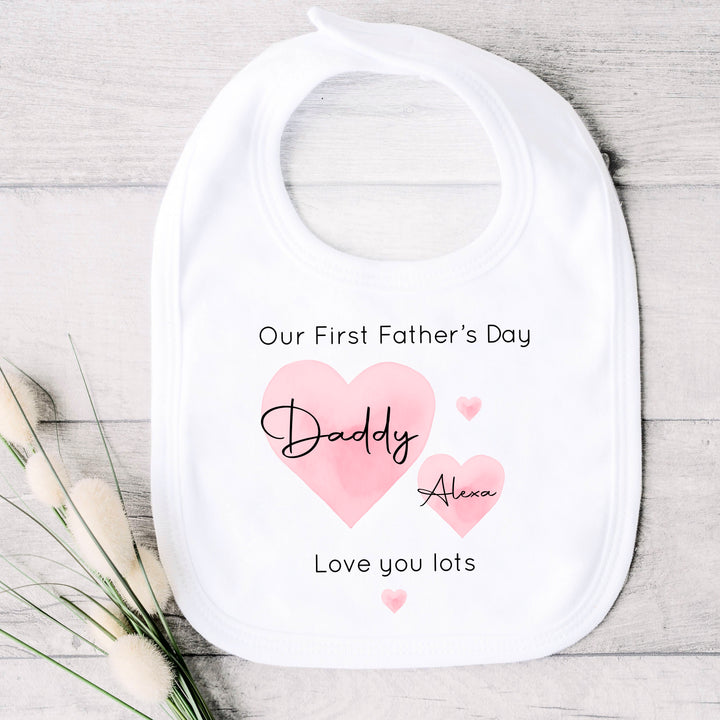 Personalised white Father's Day baby bib that says 'Our First Father's Day Daddy Alexa Love you lots'. This design features 2 big pink hearts