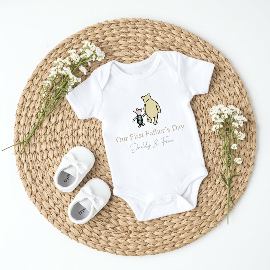 Personalised white Father's Day baby vest that says 'Our First Father's Day Daddy & Finn'. This design features the Classic Winnie the Pooh and piglet holding hands