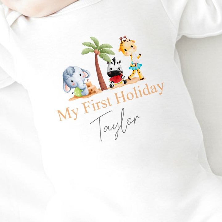 Personalised white baby grow/sleepsuit that says 'My First Holiday Taylor'. This design features 3 cartoon characters standing around a palm tree