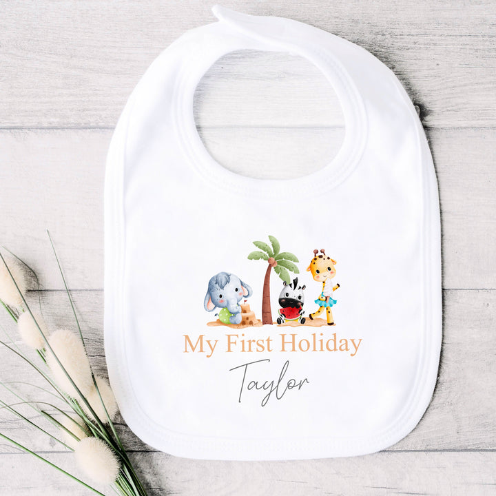 Personalised white baby bib that says 'My First Holiday Taylor'. This design features 3 cartoon characters standing around a palm tree