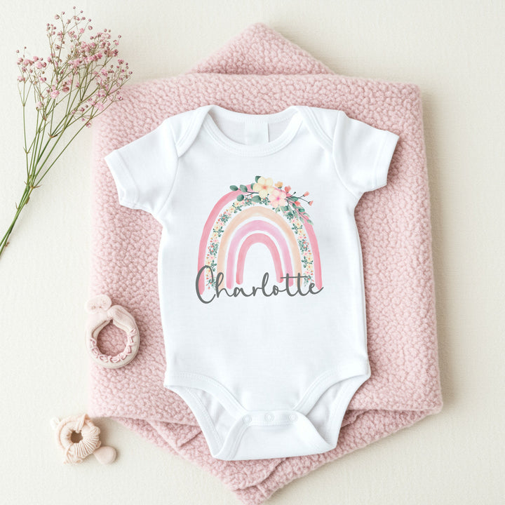 a personalised white baby vest that says 'Charlotte'. This design features a pink floral rainbow