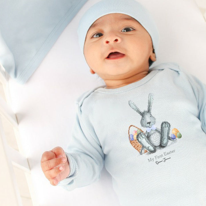 Personalised blue Easter baby grow/sleepsuit that says 'My First Easter Oscar James'. This design features a grey rabbit with blue details sitting next to an Easter basket full of eggs