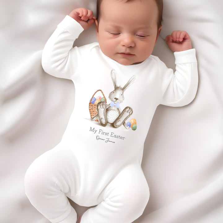 Personalised white Easter baby grow/vest that says 'My First Easer Oscar James'. This design features a grey rabbit with a bluw bow tie sitting next to an Easter basket full of eggs