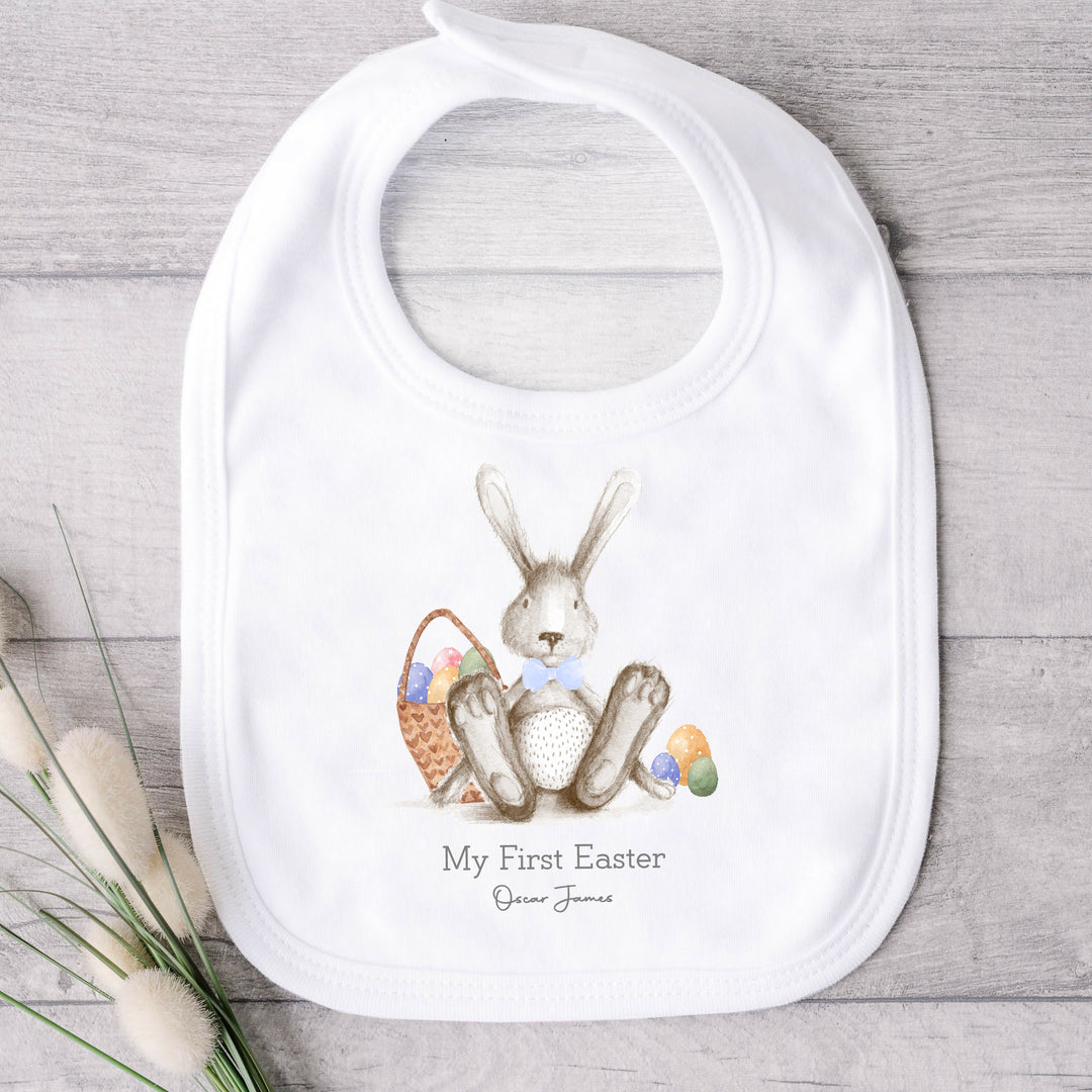 Personalised white Easter baby bib that says 'My First Easer Oscar James'. This design features a grey rabbit with a bluw bow tie sitting next to an Easter basket full of eggs