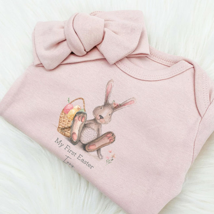 Personalised Pink Easter baby grow/sleeppsuit that says 'My First Easter Izzy'. This design features a grey rabbit with pink details sitting next to an Easter basket with eggs in it 