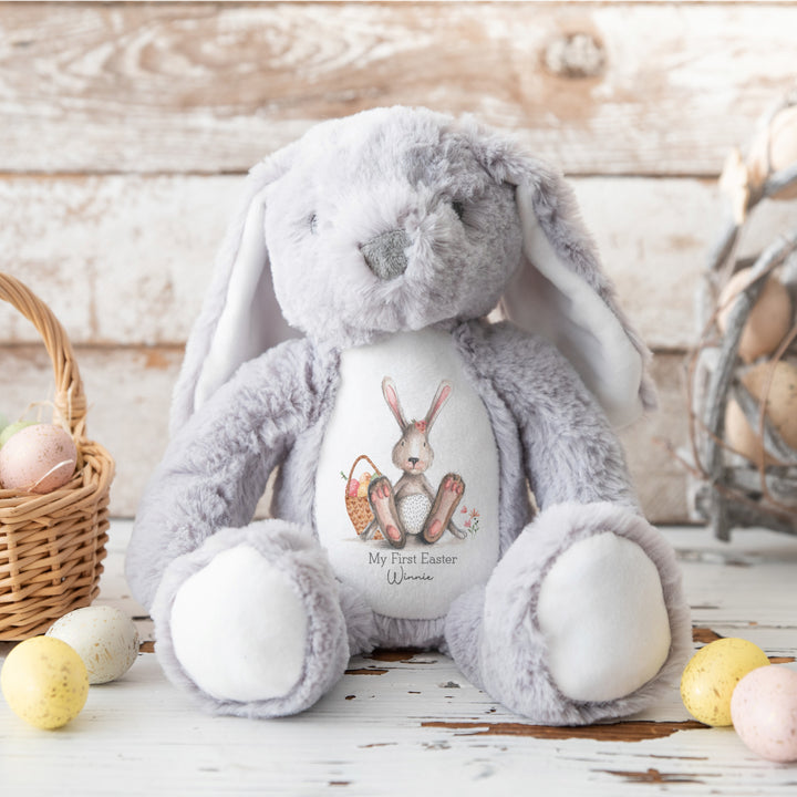 Personalised grey bunny Easter teddy that says 'My First Easter Winnie'. This design features a grey bunny with a pink bow sitting next to a basket full of Easter eggs