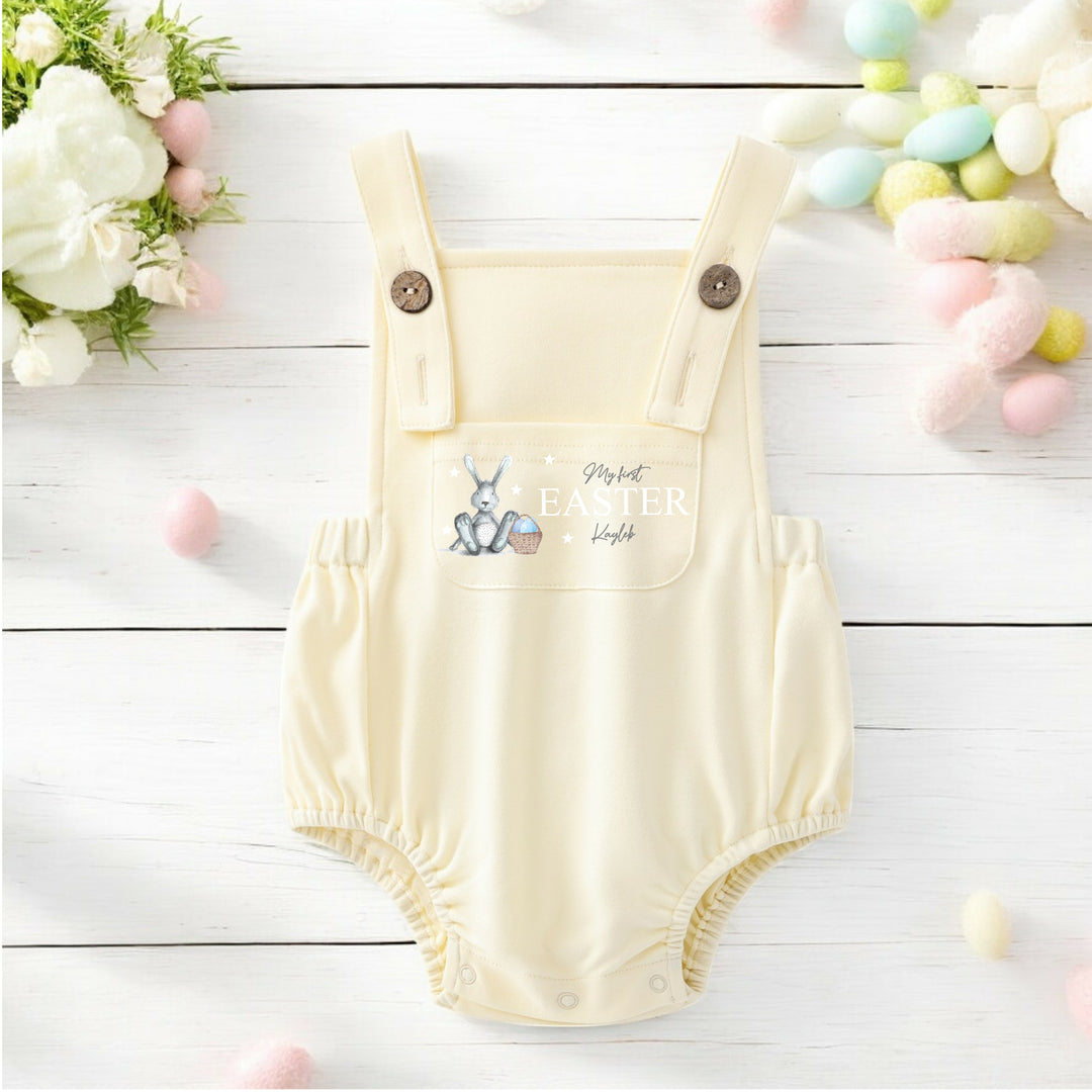 Persoanlised lemon yellow Easter dungarees that say 'My First Easter Kayleb'. This design features a grey bunny with blue details sitting next to an Easter basket full of eggs