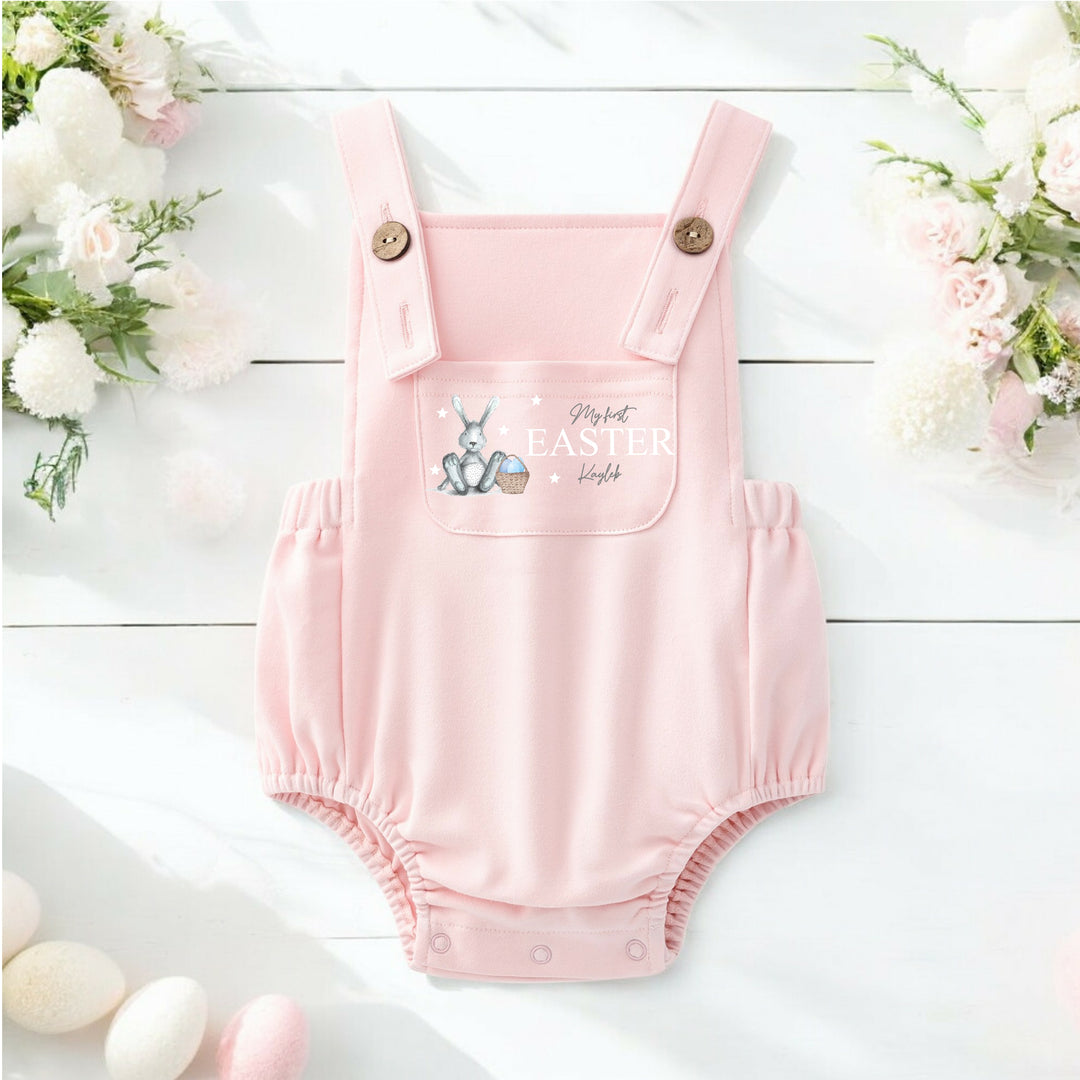 Persoanlised pink Easter dungarees that say 'My First Easter Kayleb'. This design features a grey bunny with blue details sitting next to an Easter basket full of eggs