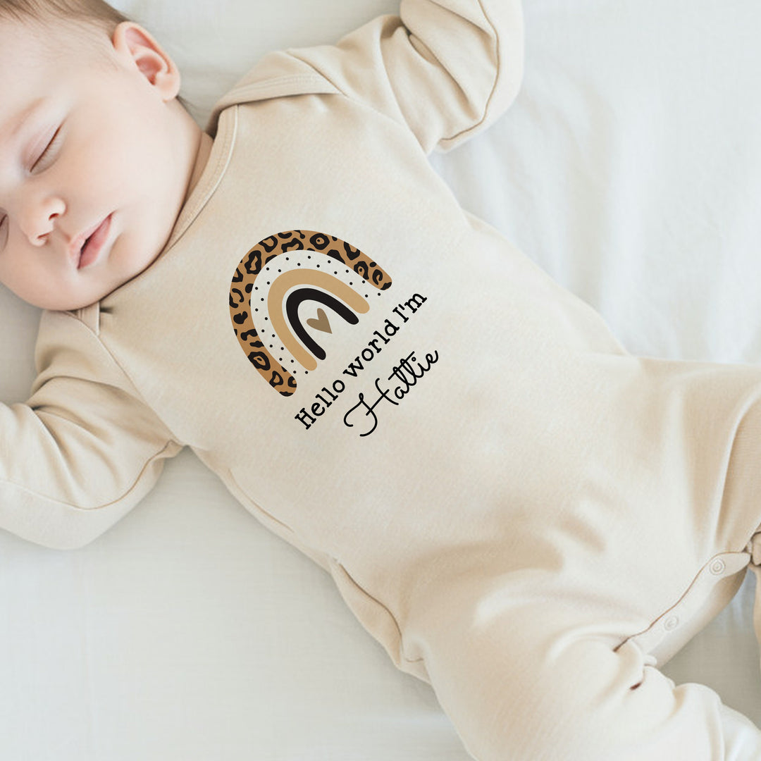 a personalised beige baby grow that says 'Hello World I'm Hattie'. This design features a leopard print rainbow