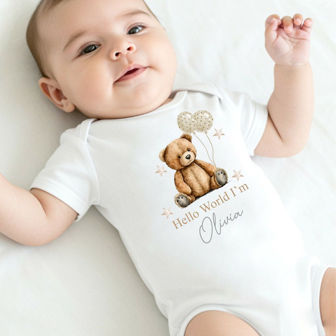A personalised white baby vest that says 'Hello World I'm Olivia'. This design features a brown teddy holding 2 beige balloons surrounded by stars