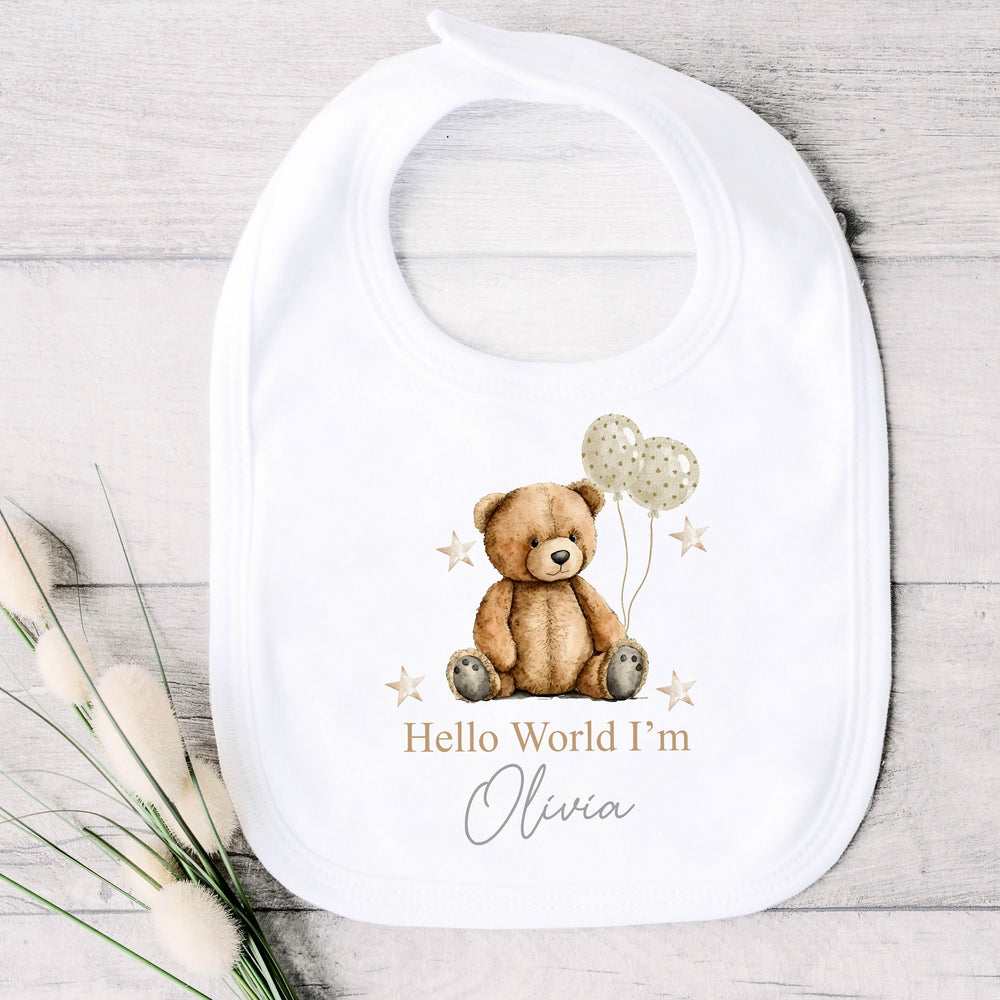 A personalised white baby bib that says 'Hello World I'm Olivia'. This design features a brown teddy holding 2 beige balloons surrounded by stars