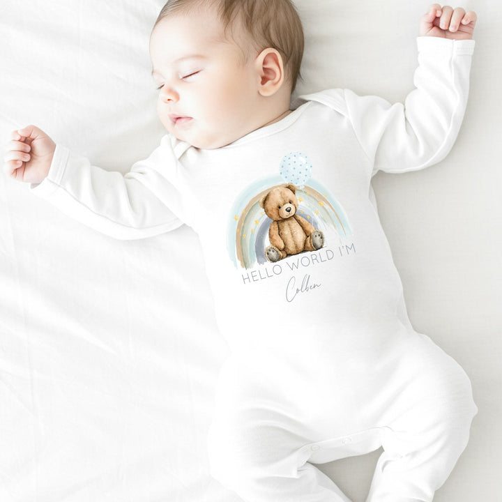 a personalised white baby grow that says 'Hello world I'm Colben'. This design features a blue rainbow with a teddy sitting in front of it holding a blue balloon