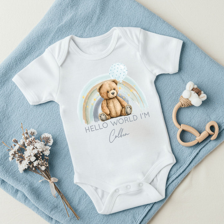a personalised white baby vest that says 'Hello world I'm Colben'. This design features a blue rainbow with a teddy sitting in front of it holding a blue balloon