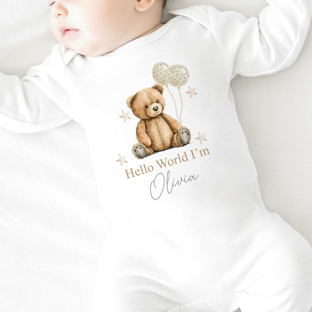A personalised white baby grow that says 'Hello World I'm Olivia'. This design features a brown teddy holding 2 beige balloons surrounded by stars