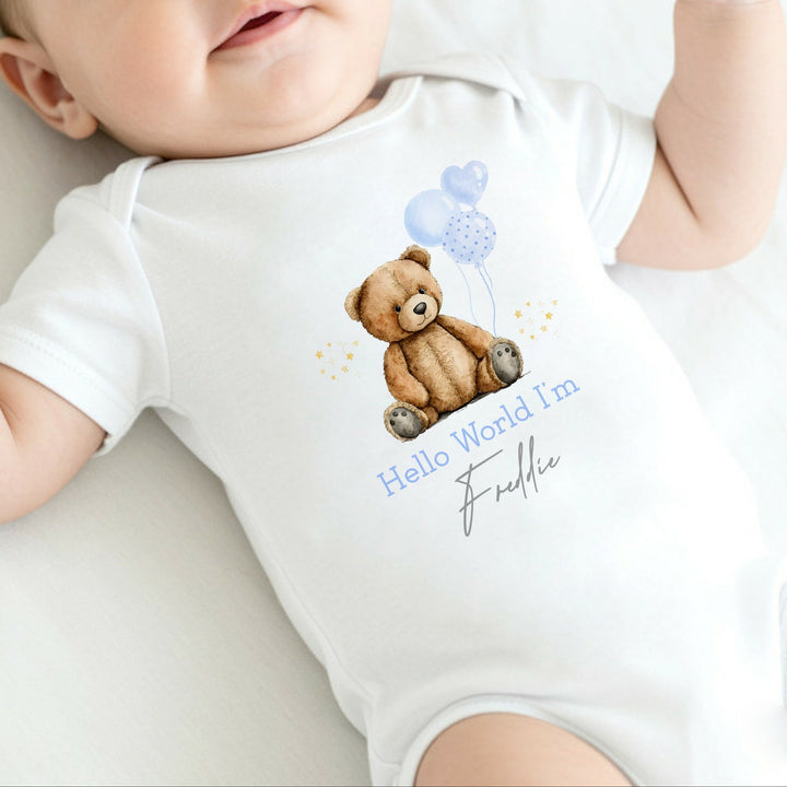 a personalised white baby vest that says 'Hello World I'm Freddie'. This design features a teddy bear holding a blue bunch of blue balloons