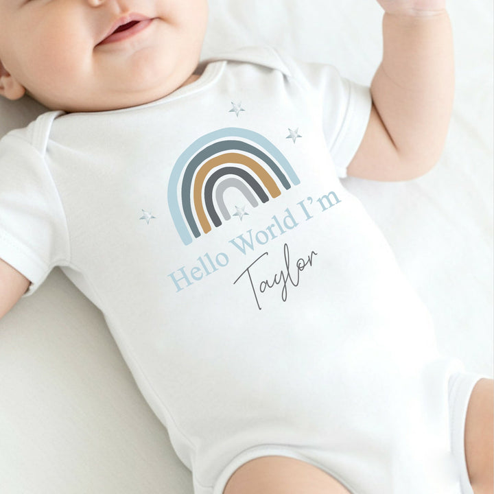 a personalised white baby vest that says 'Hello World I'm Taylor'. This design features a blue rainbow