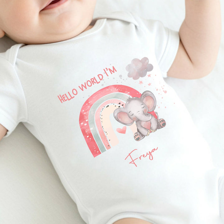 personalised white baby vest that says 'Hello world I'm Freya'. This design features a pink rainbow with an elephant sitting in front of it