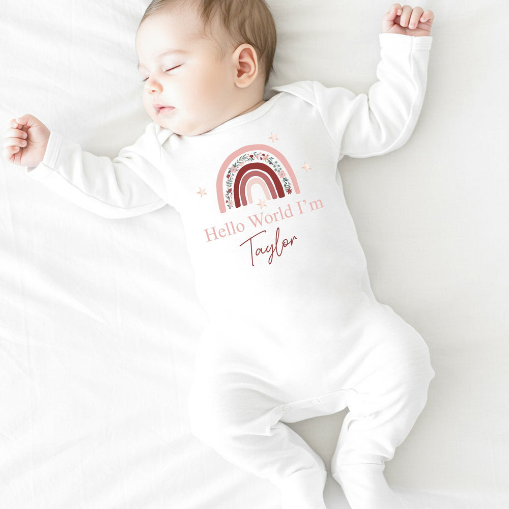 a personalised white baby grow that says 'Hello World I'm Taylor'. This design features a pink floral rainbow